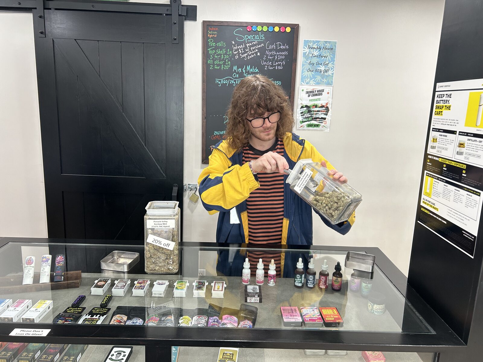 A budtender measuring flower from a glass jar with a metal scoop behind the display case